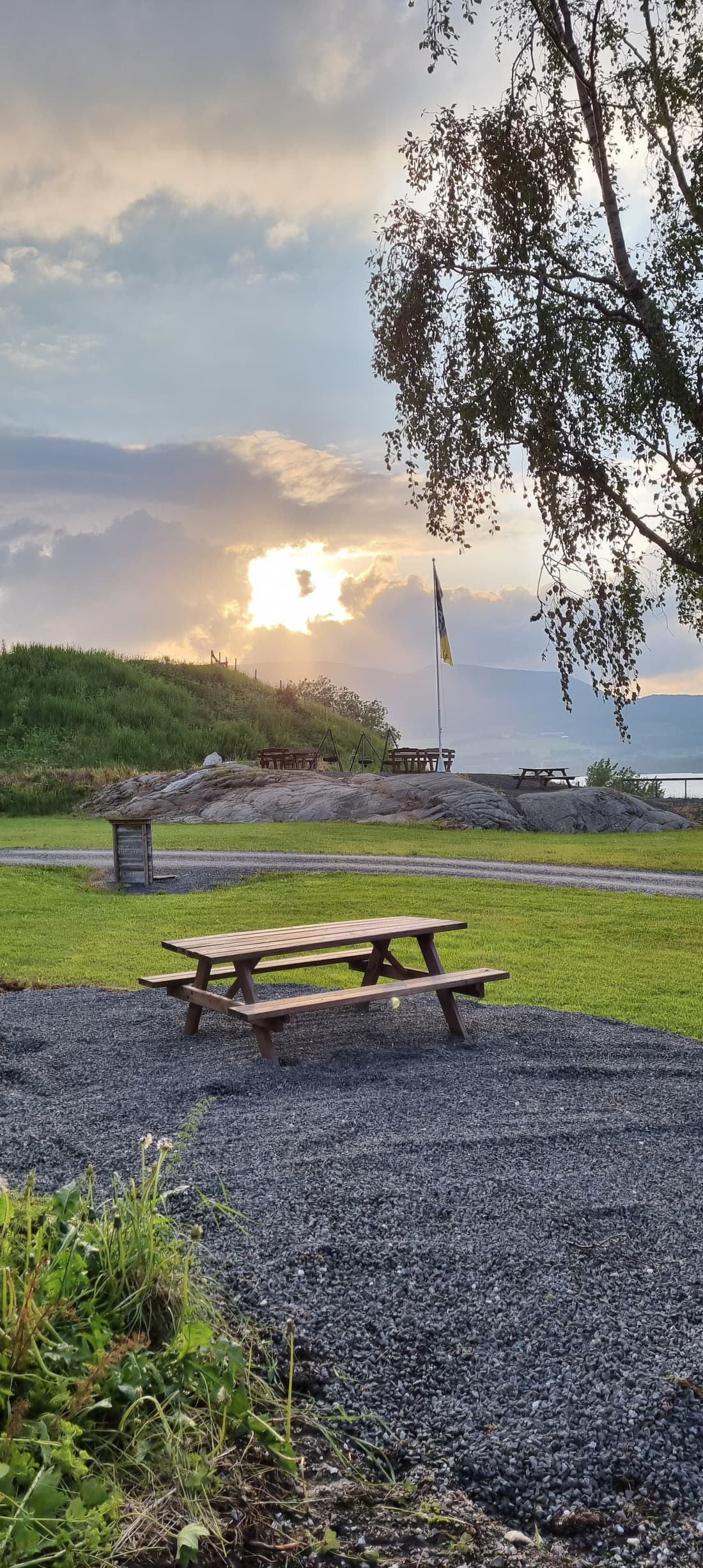 Tisch und Bänke auf dem Schotter beim Fahnenmast im Abendlicht
